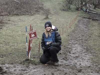 Camilla Thurlow crouches down next to a mine warning sign in a field wearing protective gear