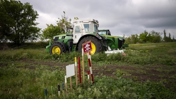 A large agricultural machine stands in the middle of a field behind a landmine warning sign.
