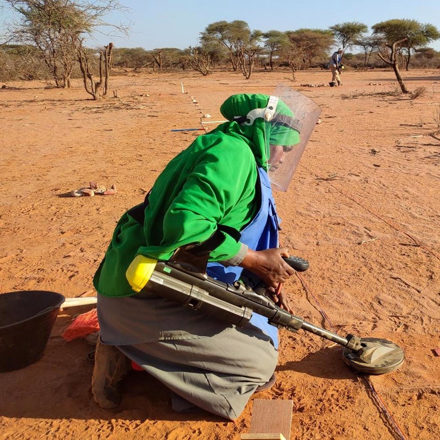 A woman in PPE uses a metal detector to carry out demining activities on ground