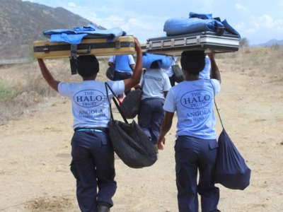 Women wearing HALO uniform in Angola carry demining kit through rural terrain