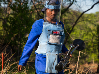 A deminer wearing protective gear walks through a field whilst holding a detector