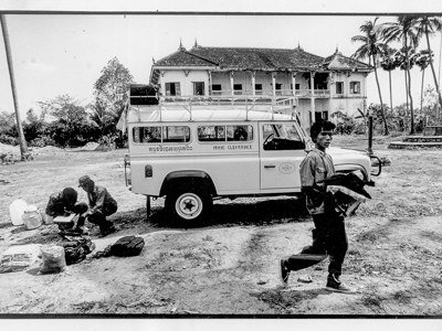 A black and white image of a HALO vehicle in front of a house in Cambodia, with HALO Trust staff 
