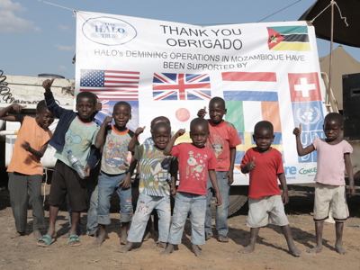 A large group of children smile and give a thumbs-up to the camera in front of a banner declaring Mozambique's mine free status