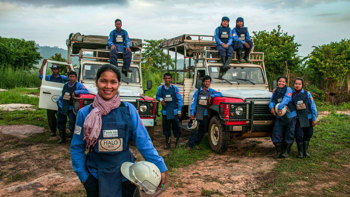 A group of deminers in Cambodia smile in front of their vehicles wearing PPE