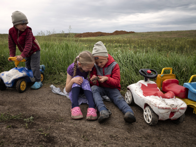 Two young children play together in a field. One boy rides a toy car to the left side.