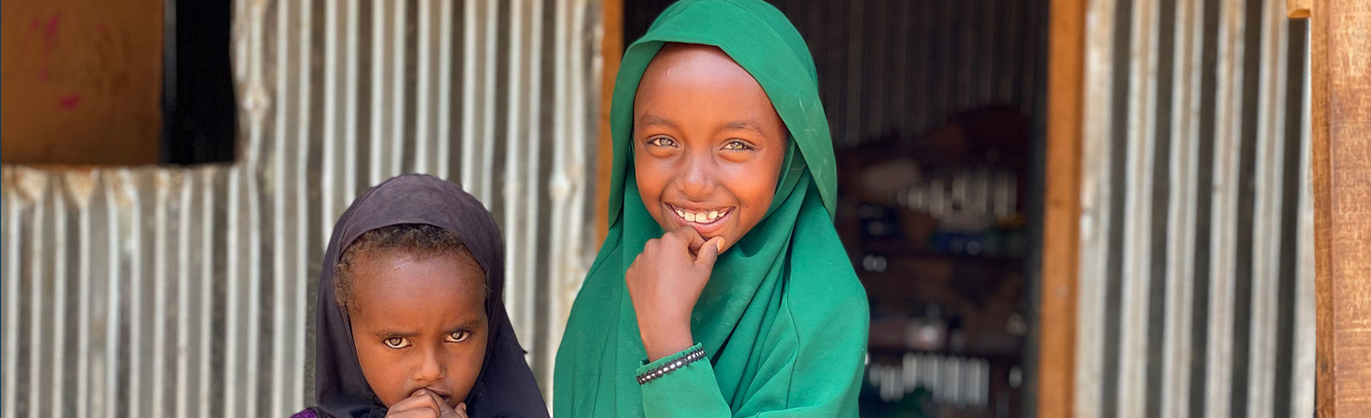 Two young girls stand outside a room made from metal sheets
