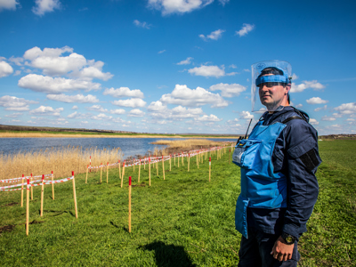 A HALO Trust deminer stands in a rural minefield next to a lake, with contamination clearly marked in front of him