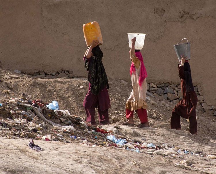 Three women walk through rubble as they carry buckets of water on their heads